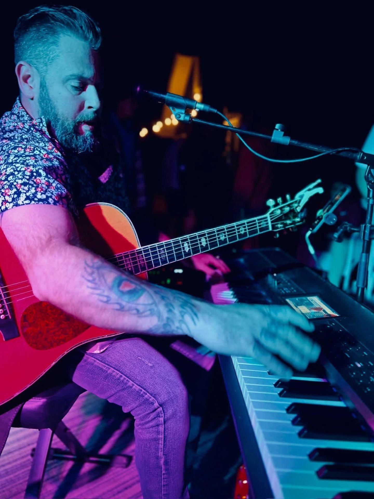Dan Signor performing seated with acoustic guitar, keyboard, and microphone under blue and magenta stage lighting