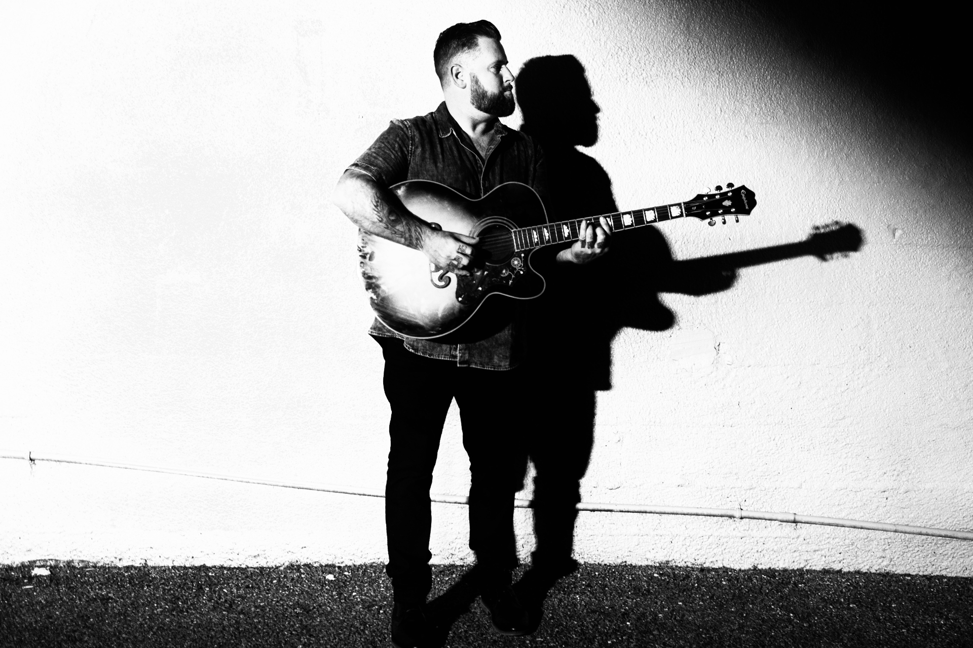 Black and white portrait of Dan with acoustic guitar against a white wall